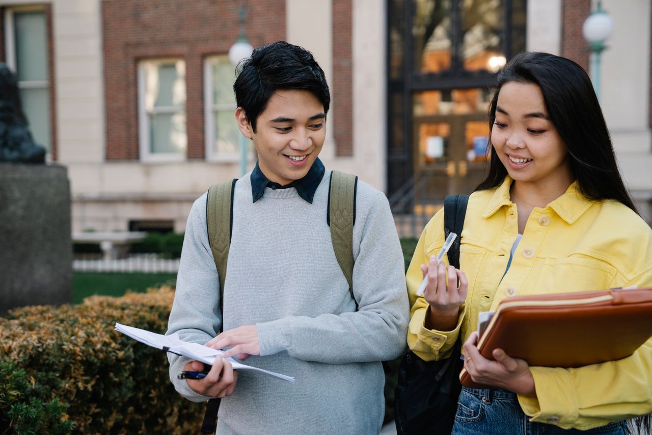 Two students collaborate outdoors on a college campus, sharing smiles and study materials.